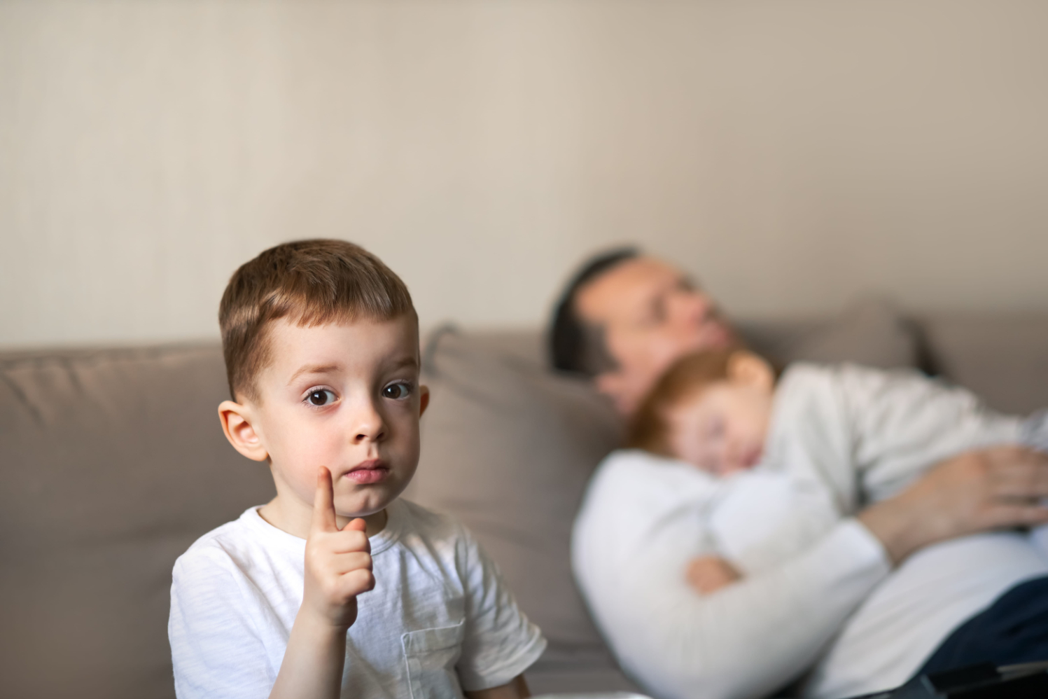 small steps Young boy in a white shirt raises a finger toward the camera, with a blurred man and child napping on a couch in the background.